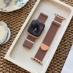 May include: A rose gold Apple Watch with a matching mesh band, displayed in a rectangular concrete tray. The watch face is black with a radial design. The band is detached, showing the clasp.