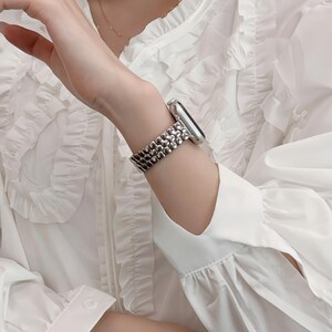 May include: A silver-toned watch with a textured band is worn on a wrist. The watch band has a beaded design. The person is wearing a white blouse with ruffled details.