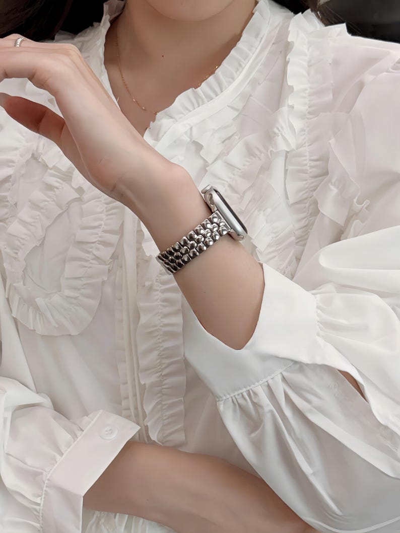 May include: A silver-toned watch with a textured band is worn on a wrist. The watch band has a beaded design. The person is wearing a white blouse with ruffled details.