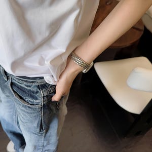 May include: A silver watch with a textured band is worn on a wrist. The person is wearing a white t-shirt and blue jeans. The jeans have a faded, distressed look. White sneakers are visible.