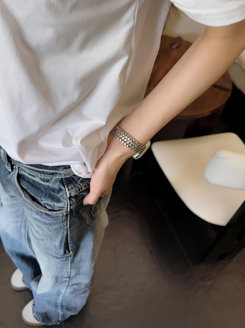 May include: A silver watch with a textured band is worn on a wrist. The person is wearing a white t-shirt and blue jeans. The jeans have a faded, distressed look. White sneakers are visible.