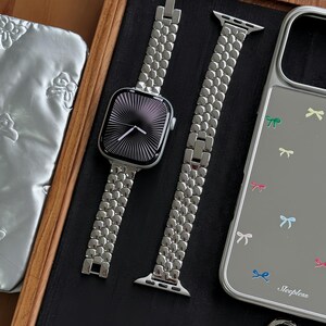 May include: A silver Apple Watch with a matching metal band, a silver phone case with bow graphics, a silver ring, and a keychain. The watch face has a black and white radial design. The phone case has the word "Sleepless" printed on it.