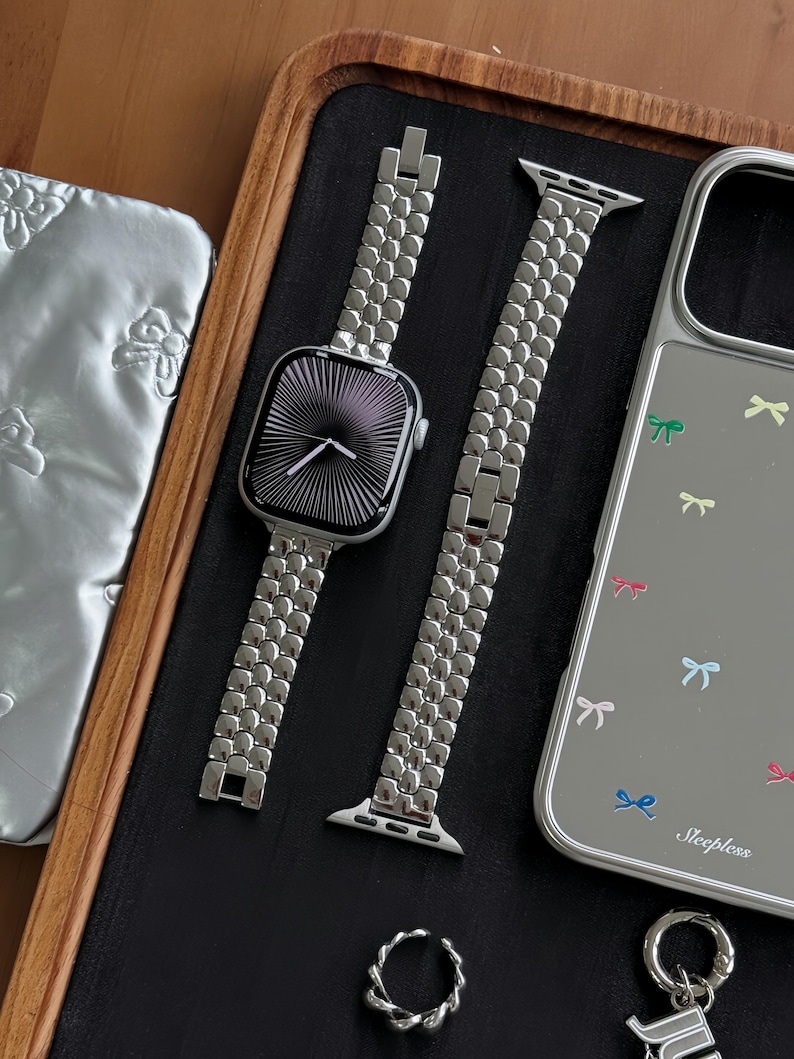 May include: A silver Apple Watch with a matching metal band, a silver phone case with bow graphics, a silver ring, and a keychain. The watch face has a black and white radial design. The phone case has the word "Sleepless" printed on it.