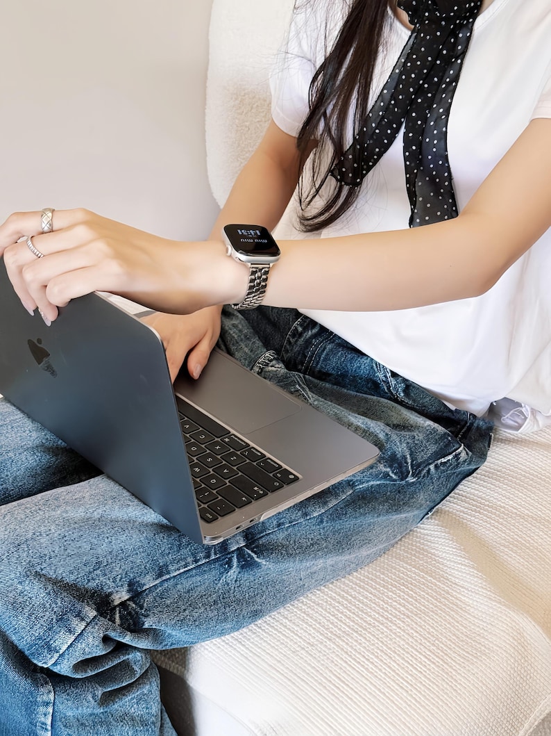 May include: A silver watch with a linked band is worn on a wrist. The watch face displays the time. The person is wearing a white t-shirt and blue jeans. A laptop is open on the person's lap.