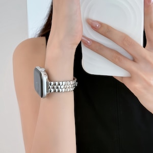 May include: A silver-toned smartwatch with a linked metal band is worn on a wrist. The watch has a rectangular face. The band is made of small, rounded links. The background is a blurred view of a person holding a white phone.