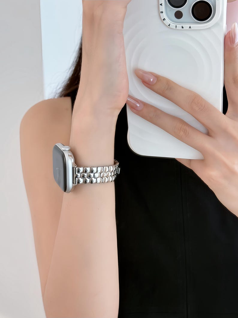 May include: A silver-toned smartwatch with a linked metal band is worn on a wrist. The watch has a rectangular face. The band is made of small, rounded links. The background is a blurred view of a person holding a white phone.