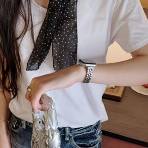 May include: A white t-shirt paired with blue jeans. A black scarf with white polka dots is draped around the neck. A silver watch with a linked band is worn on the wrist, and a silver handbag is held in the hand.