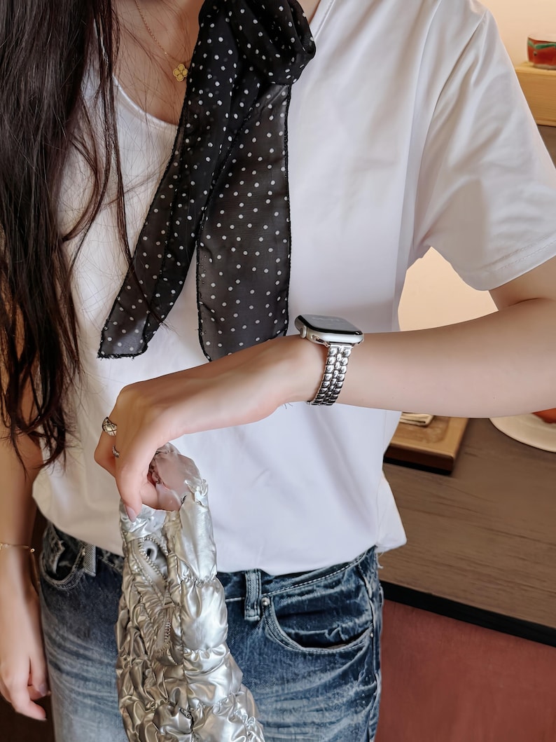 May include: A white t-shirt paired with blue jeans. A black scarf with white polka dots is draped around the neck. A silver watch with a linked band is worn on the wrist, and a silver handbag is held in the hand.
