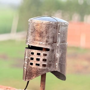 May include: A weathered, antique-style metal helmet, likely for display or costume. The helmet is a dark, metallic color with rivets and a visor featuring rectangular openings. It is mounted on a rusty metal stand, set against a blurred outdoor background.