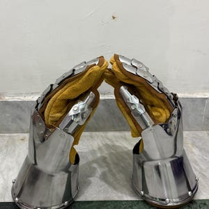 May include: Pair of medieval gauntlets. The gauntlets are made of polished steel with articulated plates and rivets. The interior is lined with yellow leather. The gauntlets are designed to protect the hands and wrists.