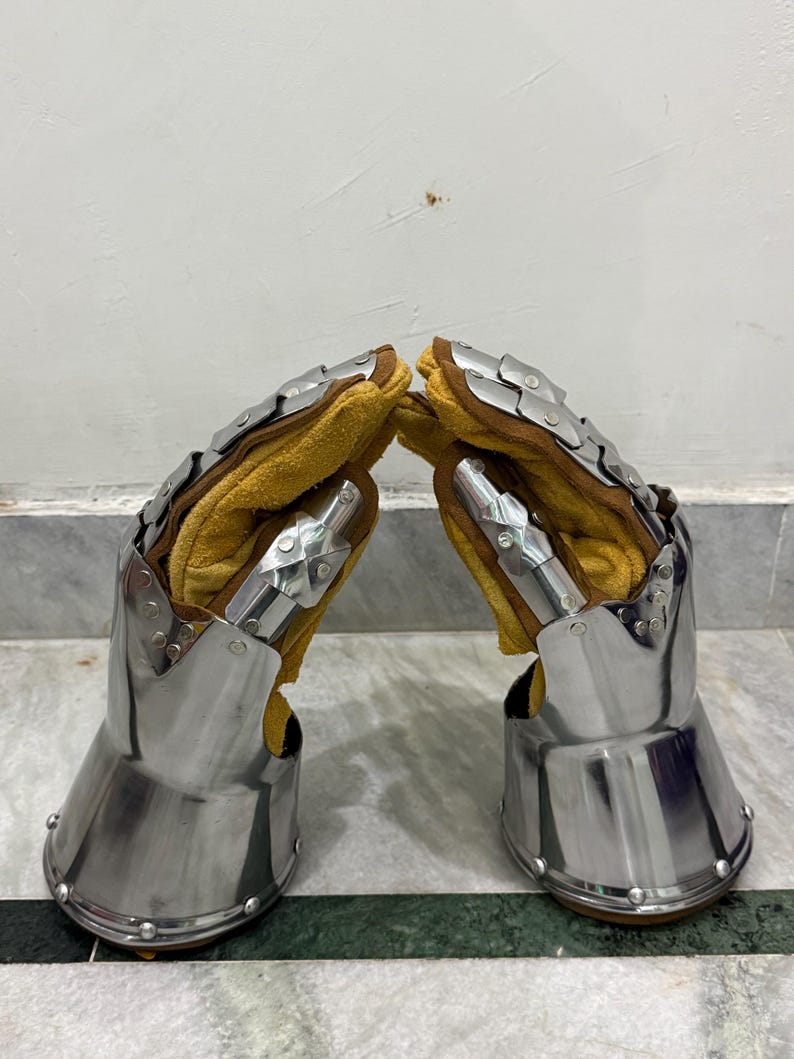 May include: Pair of medieval gauntlets. The gauntlets are made of polished steel with articulated plates and rivets. The interior is lined with yellow leather. The gauntlets are designed to protect the hands and wrists.