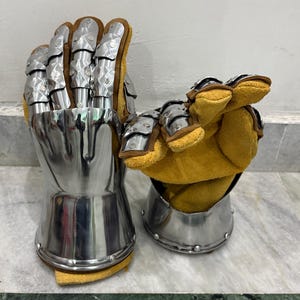 May include: Pair of medieval-style gauntlets. The gauntlets are made of polished metal with articulated finger plates and a flared cuff. The interior is lined with tan leather. The gauntlets are designed to protect the hands and wrists.