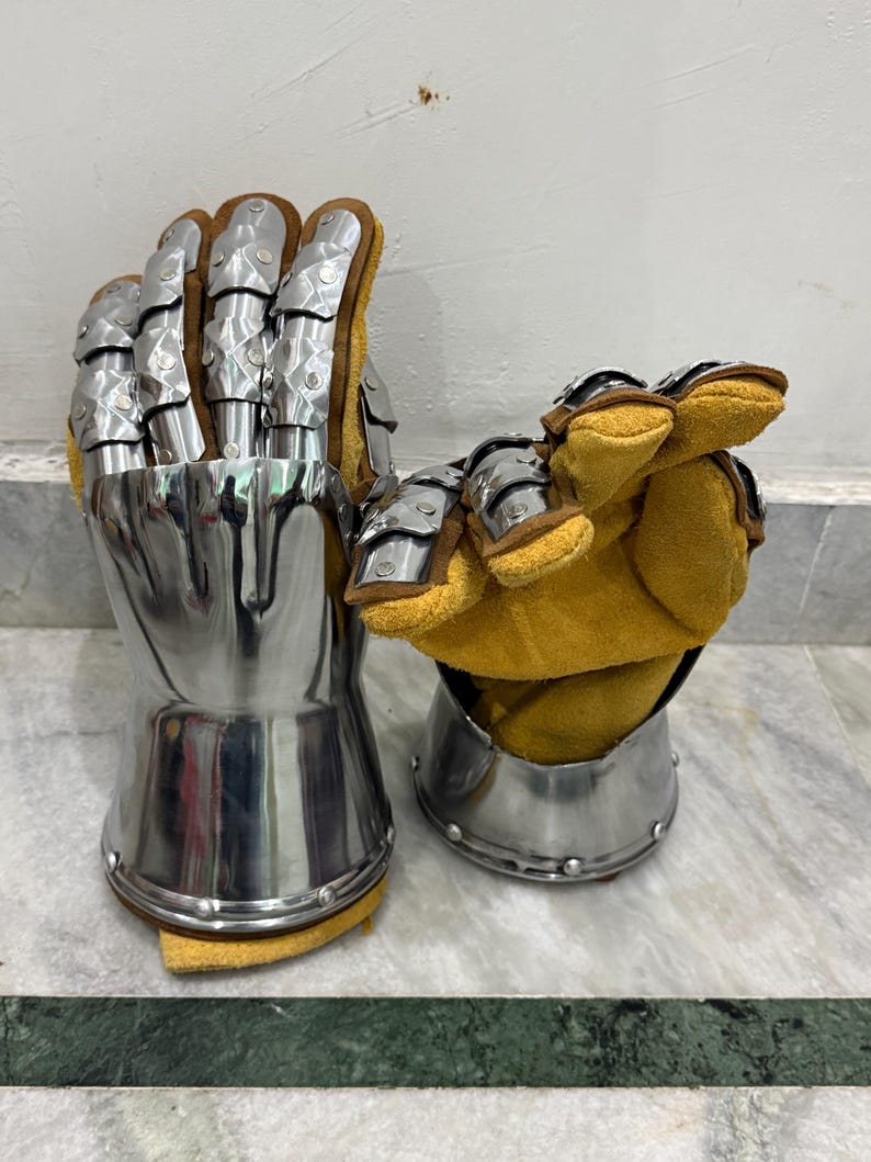 May include: Pair of medieval-style gauntlets. The gauntlets are made of polished metal with articulated finger plates and a flared cuff. The interior is lined with tan leather. The gauntlets are designed to protect the hands and wrists.