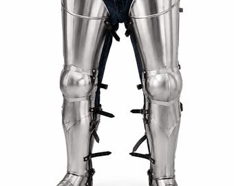May include: Polished steel leg armor covering the thighs, knees, and lower legs, featuring articulated plates and pointed sabatons. The armor is secured with black leather straps and buckles over blue denim jeans and a brown leather belt.