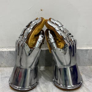 May include: Pair of medieval gauntlets made of polished steel with articulated finger plates. The interior features a tan leather lining for comfort and protection. Rivets secure the gauntlets' base.