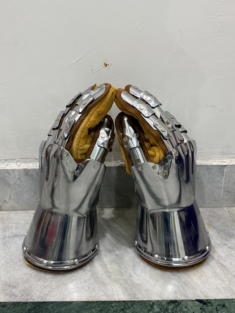 May include: Pair of medieval gauntlets made of polished steel with articulated finger plates. The interior features a tan leather lining for comfort and protection. Rivets secure the gauntlets' base.
