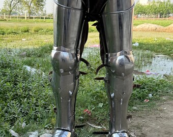May include: Polished steel leg armor, featuring articulated plates for knee and shin protection. The armor includes foot coverings and is secured with leather straps. The design is reflective and appears to be historically inspired.