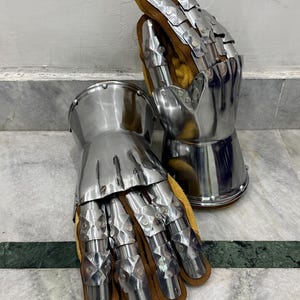May include: A pair of articulated metal gauntlets, likely for historical reenactment or display. The gauntlets feature polished steel plates and are lined with brown leather and yellow fabric. The design includes individual finger segments for flexibility.