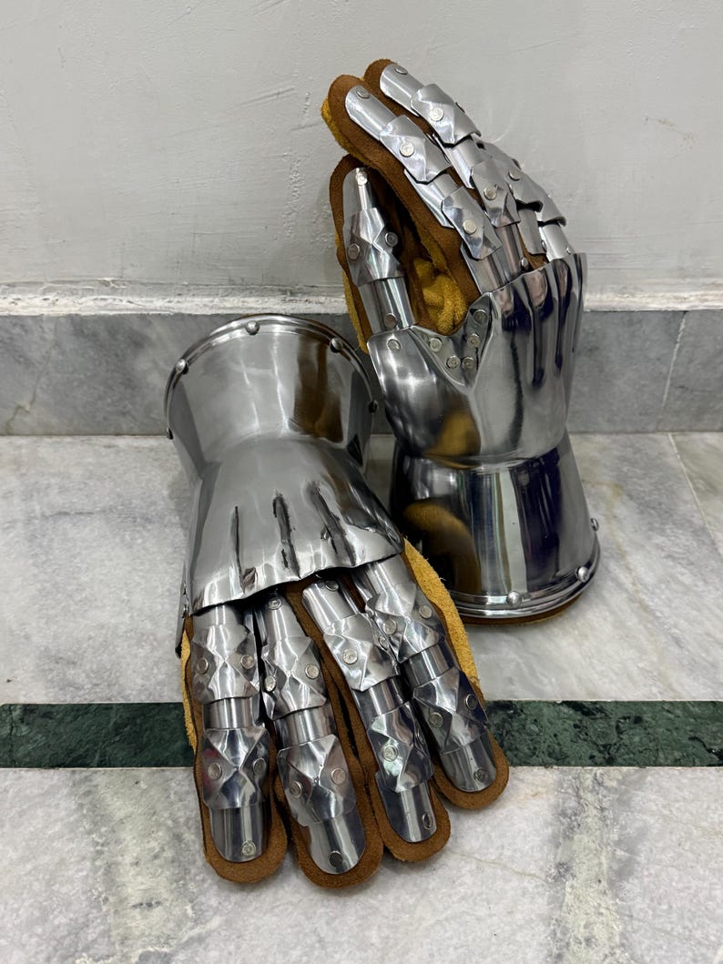 May include: A pair of articulated metal gauntlets, likely for historical reenactment or display. The gauntlets feature polished steel plates and are lined with brown leather and yellow fabric. The design includes individual finger segments for flexibility.