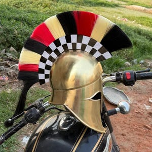 Può includere: Un casco da moto dorato con una cresta decorativa con accenti rossi, neri e gialli e un motivo a scacchi. Il casco è posizionato su una moto nera con strisce dorate.