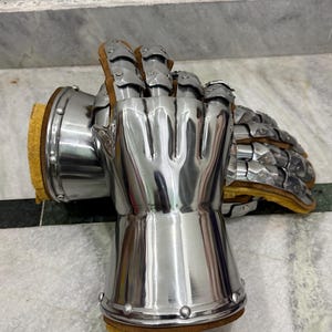 May include: Pair of polished steel gauntlets with articulated fingers and wrist protection. The gauntlets have a smooth, reflective surface and are lined with brown leather and yellow padding. Rivets secure the metal plates.