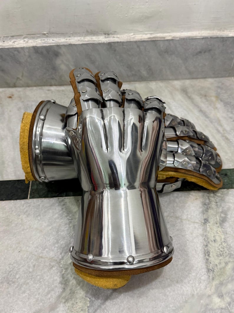 May include: Pair of polished steel gauntlets with articulated fingers and wrist protection. The gauntlets have a smooth, reflective surface and are lined with brown leather and yellow padding. Rivets secure the metal plates.