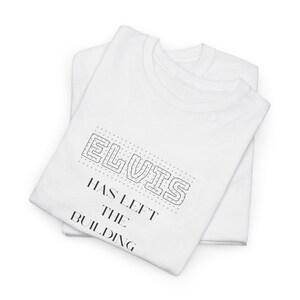 May include: Two white t-shirts are stacked, with the top shirt featuring the text "ELVIS HAS LEFT THE BUILDING" in black. The text is in a stylized font, with the word "ELVIS" outlined with dotted lines.