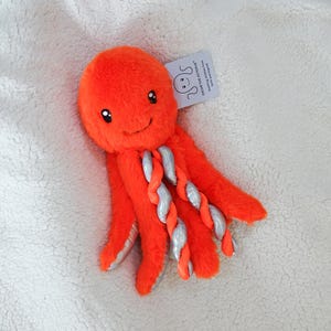 May include: A bright orange plush octopus toy with soft fur, featuring large black eyes and a small embroidered smile. Several tentacles are twisted with a shiny silver fabric underside. A white tag attached reads OSCAR THE OCTOPUS.