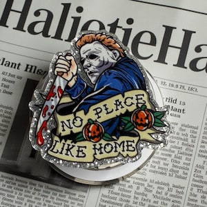 May include: A pop socket featuring a graphic of Michael Myers from the Halloween movie franchise, holding a bloody knife. The design includes the text "NO PLACE LIKE HOME" with pumpkins and roses. The pop socket is on a newspaper background.