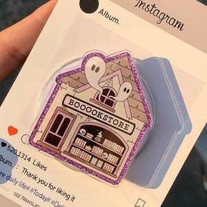 May include: A house-shaped phone grip with a bookstore design, featuring ghosts and the text "BOOOOKSTORE." The grip has a glittery purple border and is set against a social media interface.