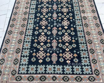 May include: A rectangular rug with a dark blue central field featuring geometric patterns in shades of pink, beige, and teal. The border has a repeating diamond and square design in similar colors. The rug has a fringe on the short ends.