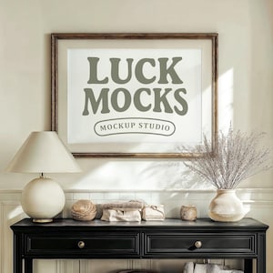 May include: A black console table with two drawers and a lower shelf, holding two woven baskets and a stack of books. A framed print with the text "LUCK MOCKS" hangs above. A lamp and vase with dried plants are on the table.