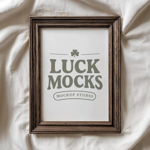 Puede incluir: Un marco de fotos rectangular de madera marrón con el texto "LUCK MOCKS" y "MOCKUP STUDIO" en gris. El marco está sobre un fondo de tela blanca.