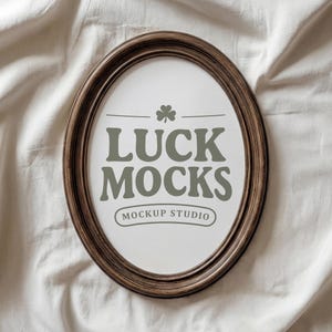 May include: An oval-shaped, brown wooden frame with the text "LUCK MOCKS" in gray, along with a shamrock graphic and "MOCKUP STUDIO" text. The frame is set against a white, textured fabric background.