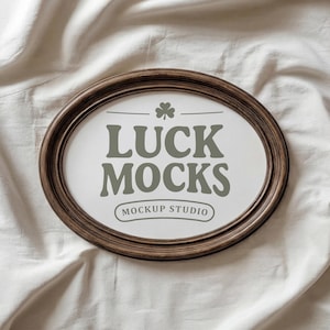 May include: Oval wooden frame with the text "LUCK MOCKS" and "MOCKUP STUDIO" in a green-grey colour. A small shamrock is above the text. The frame is on a white fabric background.