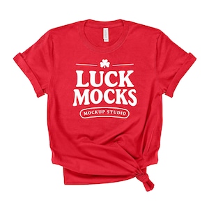 May include: A red t-shirt with the text "LUCK MOCKS MOCKUP STUDIO" in white. The shirt has a crew neck and rolled-up sleeves. The bottom of the shirt is tied in a knot. The shirt is made of a soft material.