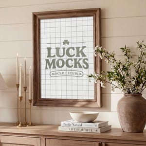 Puede incluir: Un marco de madera con el texto "LUCK MOCKS MOCKUP STUDIO" en verde claro. El marco es marrón y el fondo es un patrón de cuadrícula. Un candelabro dorado con cuatro velas está a la izquierda. Un jarrón con flores blancas está a la derecha.