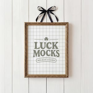 Könnte beinhalten: Ein rechteckiger, goldfarbener Rahmen mit Perlenrand, der vor einem weißen Holzhintergrund hängt. Der Rahmen enthält ein weißes Gitter mit dem grauen Text "LUCK MOCKS MOCKUP STUDIO". Oben ist ein schwarzes Band gebunden.