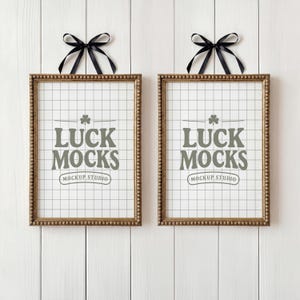 May include: Two framed prints with gold-colored borders and black ribbon bows at the top. The prints feature a grid background and the text "LUCK MOCKS MOCKUP STUDIO" in a stylized font.