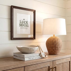 Puede incluir: Un marco con el texto "LUCK MOCKS" cuelga sobre un mueble de madera. Un cuenco de cerámica blanca y dos libros, uno titulado "THE ART OF HOME", están sobre el mueble. Una lámpara tejida con una pantalla blanca está a la derecha.