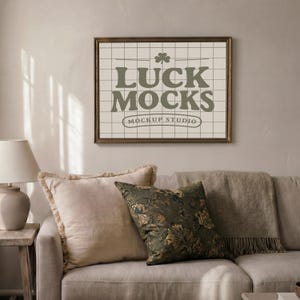 May include: A framed print with the text "LUCK MOCKS" and a shamrock graphic, displayed above a sofa with pillows and a throw blanket. The print has a grid background and a brown frame. The sofa is beige.
