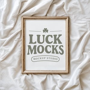 May include: A framed print with the text "LUCK MOCKS" in a bold, serif font, with a shamrock above the text. The frame is a light brown color. The words "MOCKUP STUDIO" are below the main text. The background is a white, textured fabric.