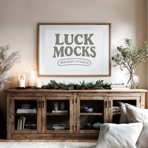 May include: A wooden cabinet with glass doors and shelves, holding books and decorative items. Above it, a framed print displays the text "LUCK MOCKS MOCKUP STUDIO". Candles and greenery add to the decor. A vase with branches and pillows complete the scene.