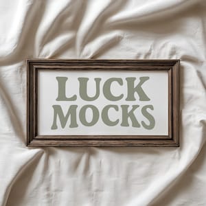 May include: A rectangular framed print with the words "LUCK MOCKS" in large, sage green letters. The frame is a dark brown color, and the background is white. The print is displayed on a white, textured surface.