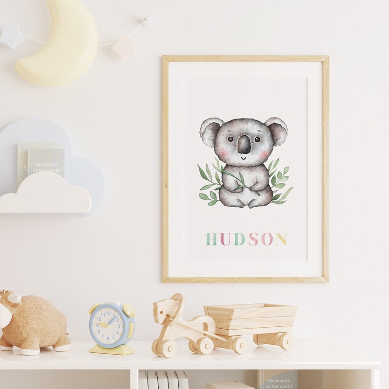 May include: A watercolour illustration of a koala bear sitting on a branch with green leaves. The name "HUDSON" is written below the koala in colourful letters. The illustration is framed in a light wood frame and is hanging on a white wall.