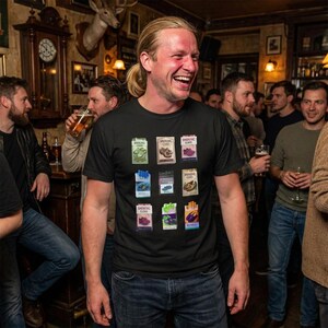 May include: Black t-shirt featuring colorful illustrations of cigarette packs. The shirt has a crew neck and is worn by a person smiling in a bar setting. The illustrations include text that reads "Smoking Kills".