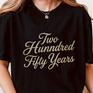 Könnte beinhalten: Schwarzes T-Shirt mit dem Text "Two Hundred Fifty Years" in goldfarbener Schreibschrift. Das Shirt hat einen Rundhalsausschnitt. Die Person trägt Armbänder und eine Halskette.
