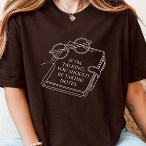 May include: A dark brown t-shirt featuring the text "IF I'M TALKING, YOU SHOULD BE TAKING NOTES." The design includes a notebook and glasses. The shirt is a relaxed fit.
