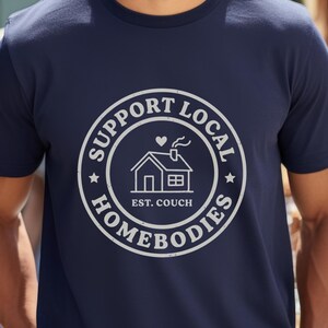 Könnte beinhalten: Marineblaues T-Shirt mit weißem, kreisförmigem Aufdruck mit der Aufschrift "SUPPORT LOCAL HOMEBODIES" um eine Hausillustration. Der Text "EST. COUCH" steht darunter. Das T-Shirt wird von einer Person mit Sonnenbrille getragen.