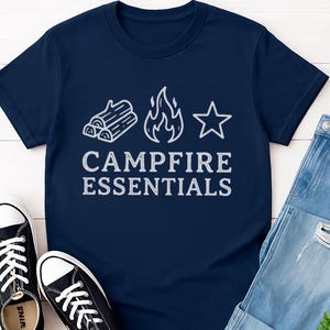 Könnte beinhalten: Marineblaues T-Shirt mit weißer Grafik einer Lagerfeuerszene und dem Schriftzug "CAMPFIRE ESSENTIALS". Das Bild zeigt auch blaue Jeans und schwarze Leinenschuhe. Eine kleine grüne Pflanze ist ebenfalls zu sehen.